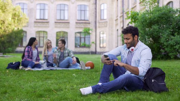 Hispanic male student sitting on grass and using cellphone, app for acquaintance - Foto, afbeelding