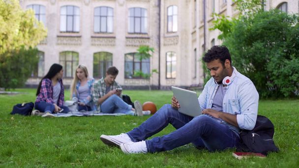 Mixed race student using laptop, sitting on grass on campus, education online - Foto, afbeelding