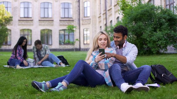 Jong (echt) paar zittend op gazon op de campus en het kijken naar foto's op smartphone - Foto, afbeelding