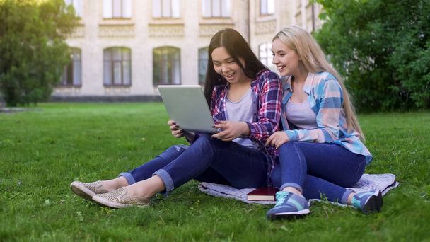 Two female students sitting on grass, laughing at pictures in laptop, shopping - Foto, immagini