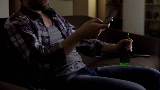 Male holding bottle of beer on knee, switching TV channels with remote control - Фото, зображення