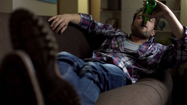 Wasted guy lying on sofa, drinking up beer from bottle, dozing off, alcoholism - Фото, зображення