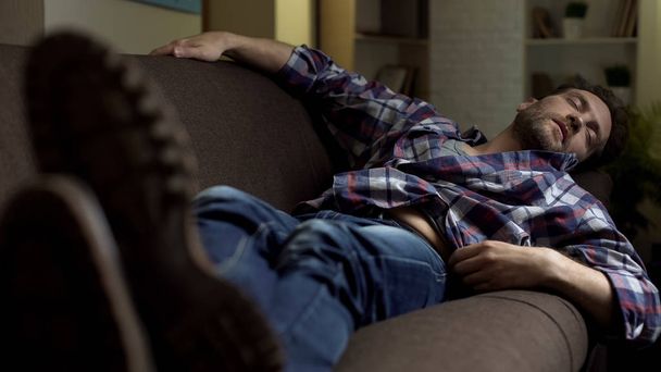 Drunken male sleeping on sofa, passive life of lazy bachelor, unemployed loser - Fotó, kép
