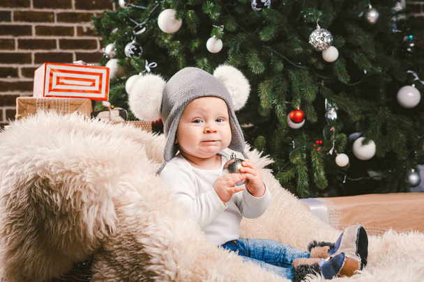 Subject children christmas new year. Caucasian little funny baby boy 1 year old sitting sleigh bear skin Christmas tree head warm hat with balabons balls, balls and jeans around box gifts in evening. - Foto, Imagen