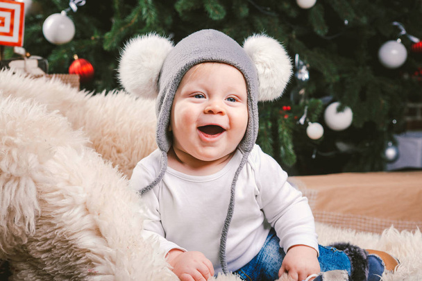 Subject children christmas new year. Caucasian little funny baby boy 1 year old sitting sleigh bear skin Christmas tree head warm hat with balabons balls, balls and jeans around box gifts in evening. - Photo, image