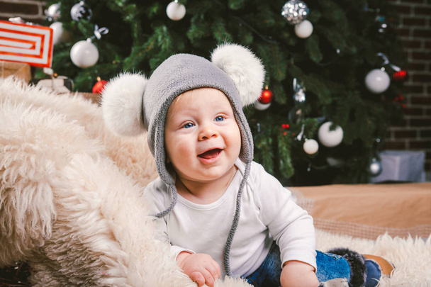 Subject children christmas new year. Caucasian little funny baby boy 1 year old sitting sleigh bear skin Christmas tree head warm hat with balabons balls, balls and jeans around box gifts in evening. - Photo, Image
