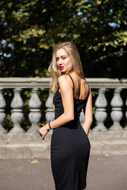 Buiten schot van verleidelijke blonde model met weelderige haar dragen van zwarte skinny jurk - Foto, afbeelding