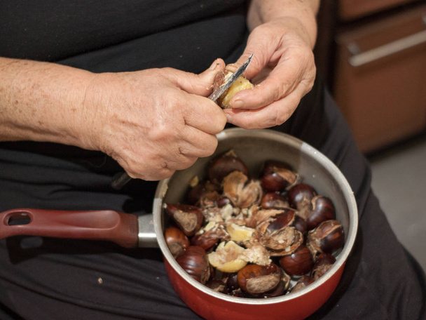 Manos de una anciana sosteniendo una olla con castañas y pelándolas con un cuchillo
 - Foto, imagen