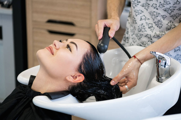 Unrecognizable professional hairdresser washing hair to her client. Beautiful hairdresser washing hair to her client lady in hairdressing saloon. Client sitting with her eyes closed. - Photo, Image