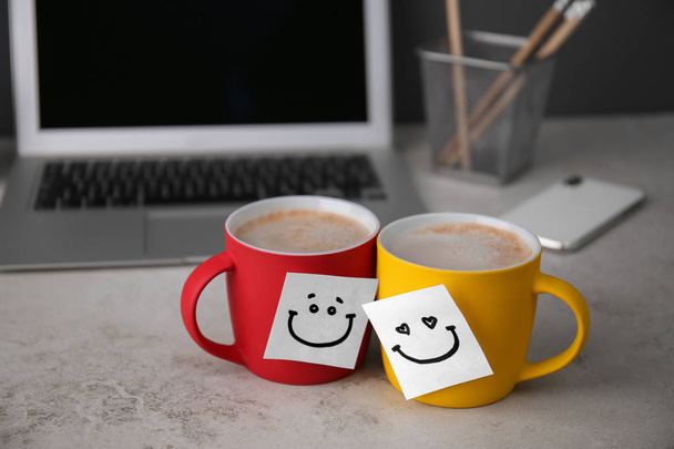 Sticky notes with funny faces attached to cups of coffee on office table. Break time - Photo, Image