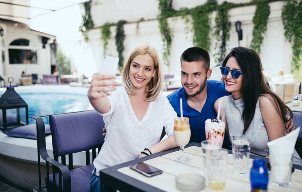 Group of young and attractive three people enjoying private party and making selfie photo. Swimming pool in background. Man with two beautiful women. - Foto, imagen