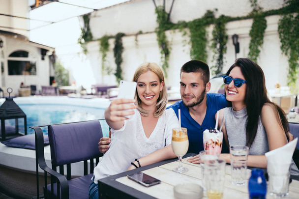 Group of young and attractive three people enjoying private party and making selfie photo. Swimming pool in background. Man with two beautiful women. - Zdjęcie, obraz