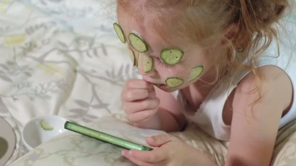 Egy gyönyörű kislány vidáman teszi egy uborkát maszkok otthon, és használ egy smartphone - Felvétel, videó