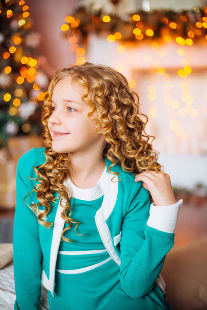 Cute little girl with curly blond hair at home near a Christmas tree with gifts and garlands and a decorated fireplace sitting on plaids and pillows - Fotó, kép