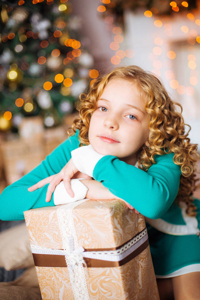 Cute little girl with curly blond hair at home near a Christmas tree with gifts and garlands and a decorated fireplace  - Valokuva, kuva