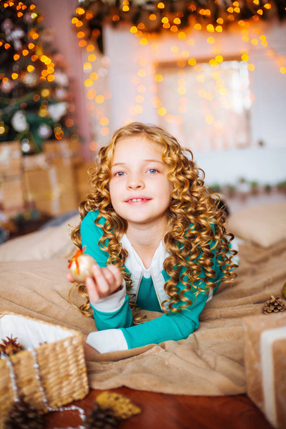 Cute little girl with curly blond hair at home near a Christmas tree with gifts and garlands and a decorated fireplace  - Fotó, kép