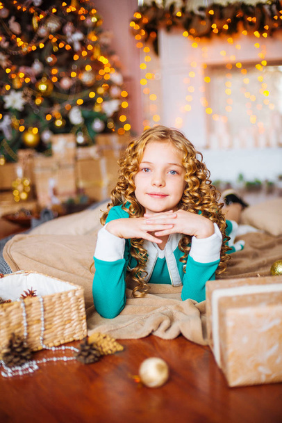 Cute little girl with curly blond hair at home near a Christmas tree with gifts and garlands and a decorated fireplace  - Фото, изображение
