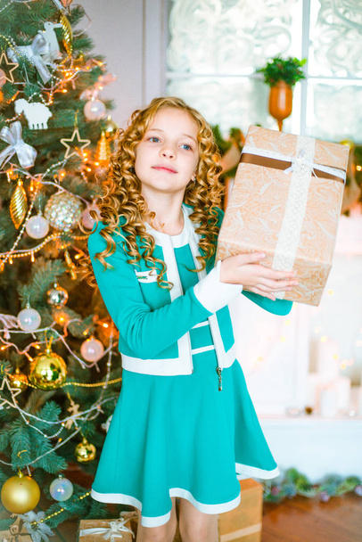 Cute little girl with curly blond hair at home near a Christmas tree with gifts and garlands and a decorated fireplace sitting on plaids and pillows - 写真・画像