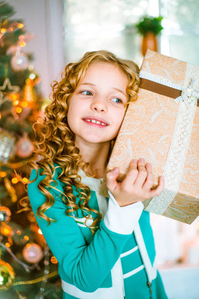 Cute little girl with curly blond hair at home near a Christmas tree with gifts and garlands and a decorated fireplace sitting on plaids and pillows - Foto, Imagem