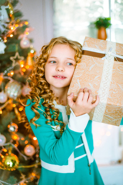 Cute little girl with curly blond hair at home near a Christmas tree with gifts and garlands and a decorated fireplace sitting on plaids and pillows - Zdjęcie, obraz