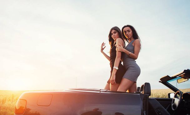 Twee meisjes graag poseren naast een zwarte auto tegen de hemel op een fantastische zonsondergang. - Foto, afbeelding