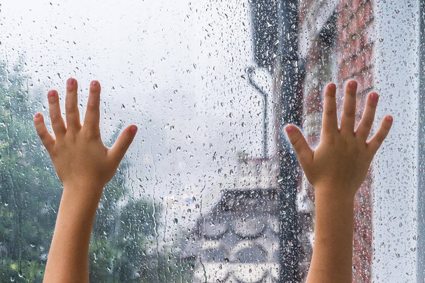 As mãos de crianças em um copo molhado com baixas. A criança do lado de fora da janela olha para a chuva de outono
 - Foto, Imagem