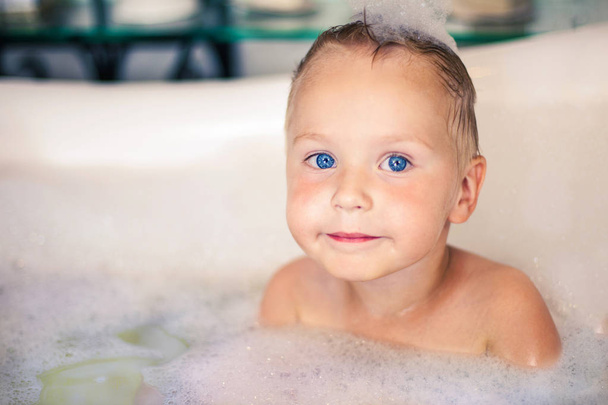 Cute little caucasian boy with bright blue eyes and blonde hair sits in the bathtube, with foam bubbles on the head. Expressive look. Indoors, copy space. - Fotó, kép