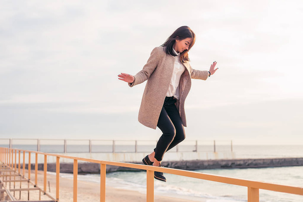 outdoor portrait of stylish brunette woman wearing grey coat jumping from railings on sea background, full length  - Фото, зображення
