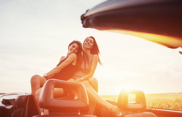 Молодые две женщины на фотосессии. Девушки с удовольствием позируют рядом с черной машиной на фоне неба на фантастическом закате
. - Фото, изображение