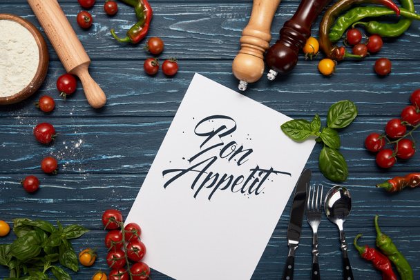 top view of template with "bon appetit" lettering, fresh vegetables, cutlery and spices on wooden surface - Фото, изображение