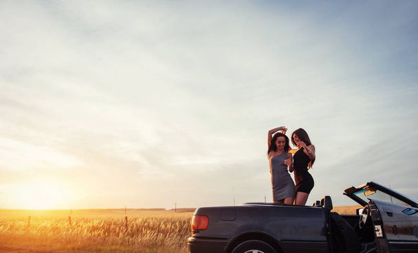 Young two women at a photo shoot. Girls gladly posing next to a black car against the sky on a fantastic sunset. - Foto, Imagem