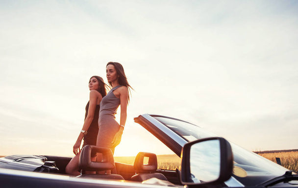 Duas jovens mulheres numa sessão fotográfica. Meninas alegremente posando ao lado de um carro preto contra o céu em um pôr do sol fantástico
. - Foto, Imagem