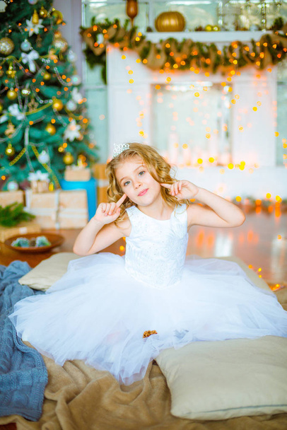 Cute little girl with curly blond hair at home near a Christmas tree with gifts and garlands and a decorated fireplace sitting on plaids and pillows - Фото, изображение