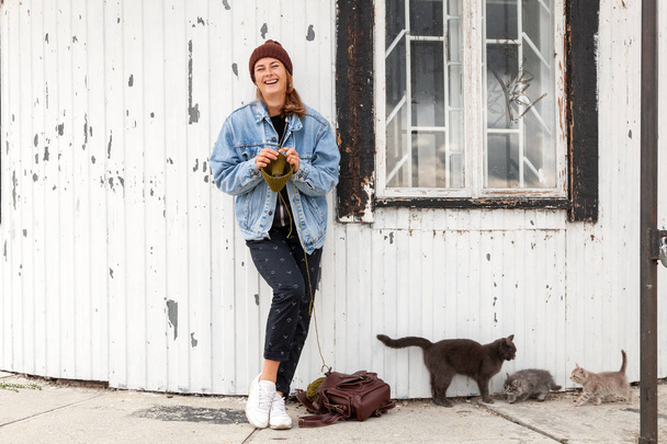Portrait young  woman  in stylish clothes knitting  green  natural wool hat, next to a cat walks with her kittens  against the background of the old lighthouse  in sunny autumn day . Freelance creative working and living concept - Foto, imagen