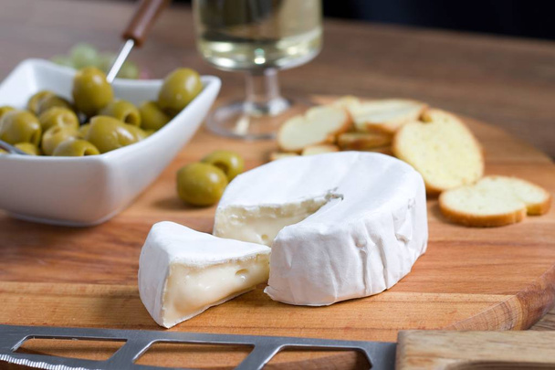 Fehér héjú lágy sajt (camembert vagy brie) fából készült táblán pirított kenyérszelettel, olajbogyóval és fehérborral - Fotó, kép