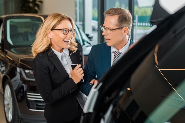 businessman and smiling businesswoman with smartphone choosing new car in dealership salon - Fotografie, Obrázek