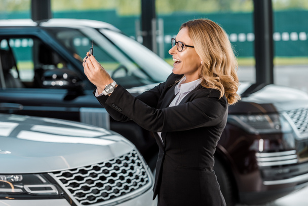 happy businesswoman in formal wear holding car key from new car in dealership salon - Photo, Image