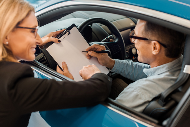 mutlu yetişkin adam arabanın içinde otururken kadın araba satıcısı ile sözleşme imzalama - Fotoğraf, Görsel