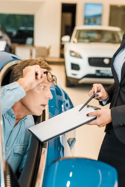 bijgesneden schot van vrouwelijke autodealer autobedrijf tonen contract aan geschokt volwassen man in de salon - Foto, afbeelding