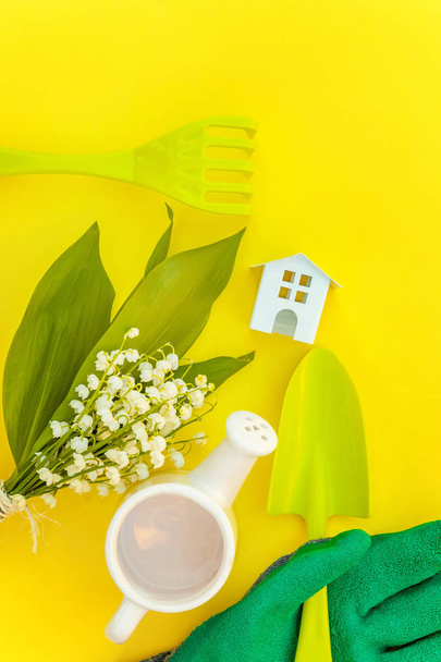 Flat Lay gardening tools, watering can, shovel, rake, glove, flowers and toy house on yellow colourful trendy background. Spring or summer in garden, eco, nature, farm, horticulture hobby concept - Photo, Image