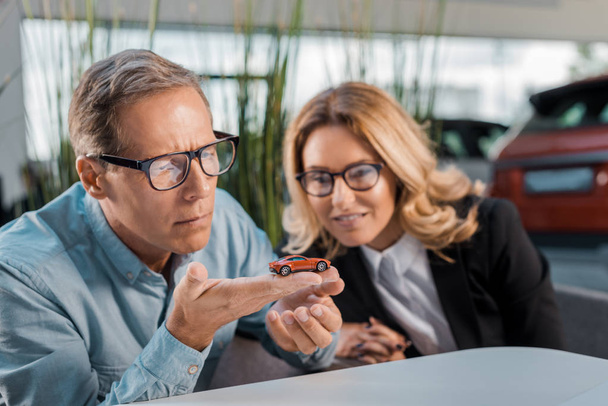 thoughtful adult customer and smiling female car dealer looking at toy car in showroom - Foto, Imagem