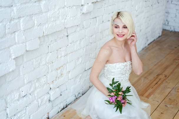 Beautiful young bride with bouquet sitting in studio on floor near white brick wall - 写真・画像