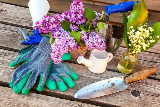Gardening tools, watering can, shovel, spade, pruner, rake, gloves, lilac, lily of the valley flowers on vintage wooden table. Spring or summer in garden, eco, nature, horticulture hobby concept - Photo, Image
