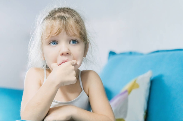 Little blonde girl sitting on the blue couch with a sad face - Фото, зображення