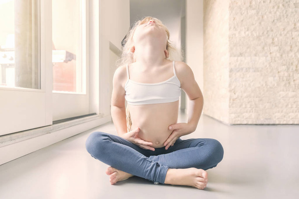 Niña rubia en top blanco y polainas azules haciendo un ejercicio de yoga en la habitación soleada
 - Foto, imagen