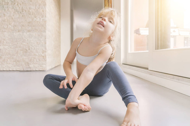Little blond girl in white top and blue leggings doing gymnastics in the sunny room - Foto, afbeelding