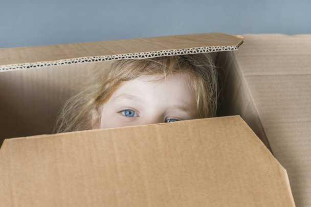 Sad little blue-eyed blonde girl looking out of a cardboard box, close-up - Foto, afbeelding