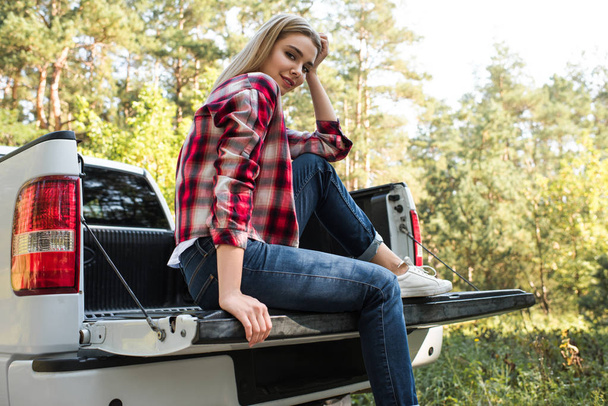 low angle view of smiling woman looking at camera and sitting in trunk of pick up car outdoors  - Fotoğraf, Görsel