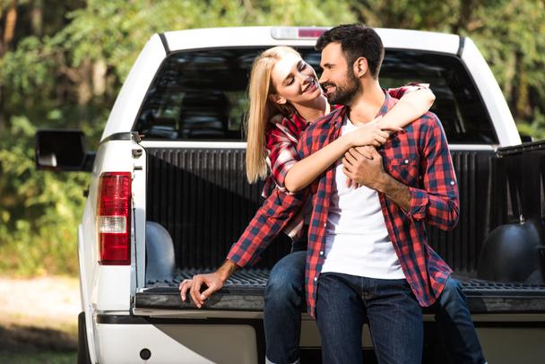young cheerful couple embracing each other on car trunk outdoors  - Photo, Image