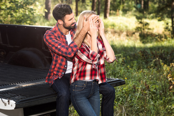 usmívající se muž pokrývající oči přítelkyni zezadu při posezení na venkovní kufru auta  - Fotografie, Obrázek